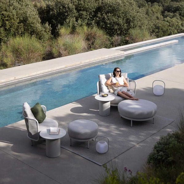 Image of woman sat on Baza modern garden lounge chair, surrounded by Baza round garden poufs and Branta circular outdoor low tables, shown on shady poolside
