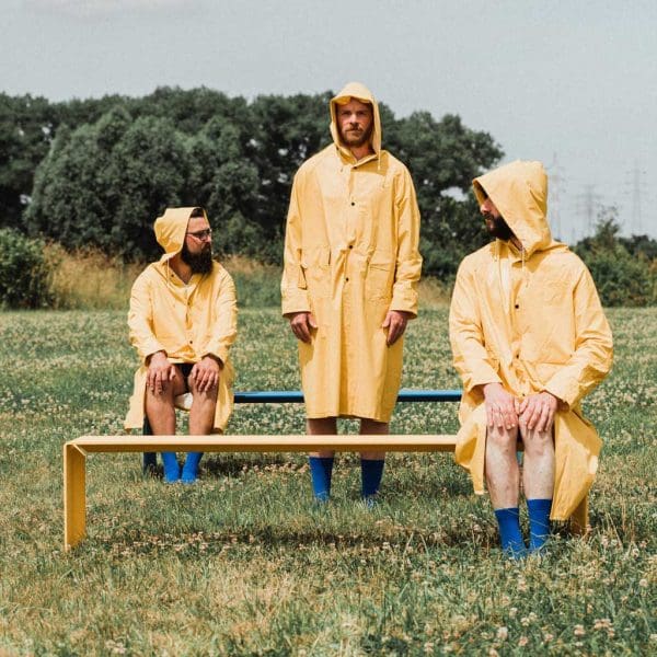 Strange image of 3 men dressed on waterproof coats, stood and sat around Wünder's The Bended modern garden benches, shown in sunny field