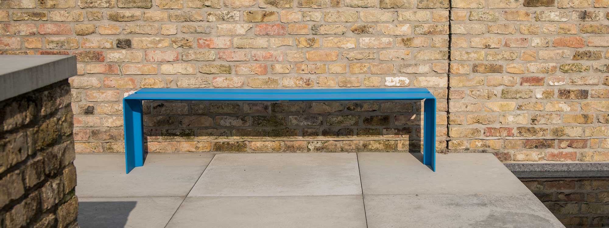 Image of Wünder's The Bended modern garden bench seat in Blue RAL 5012 aluminium, with brick wall in the background
