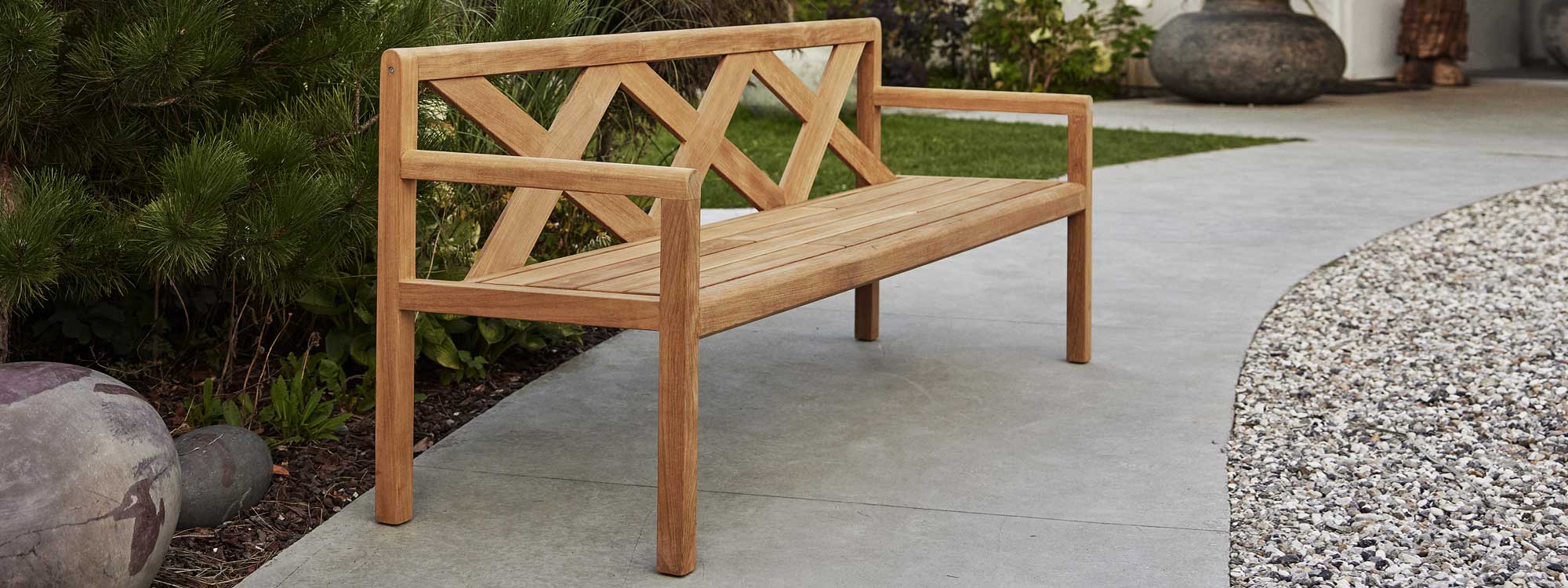Image of Grace 3 seat teak bench by Cane-line, on poured concrete pathway in garden