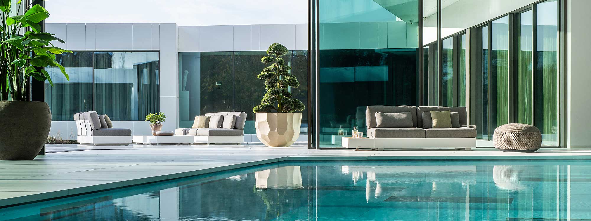 Image of Bari linear garden sofa in white aluminium with grey cushions, on indoor poolside next to large planter with cloud pruned Japanese holly