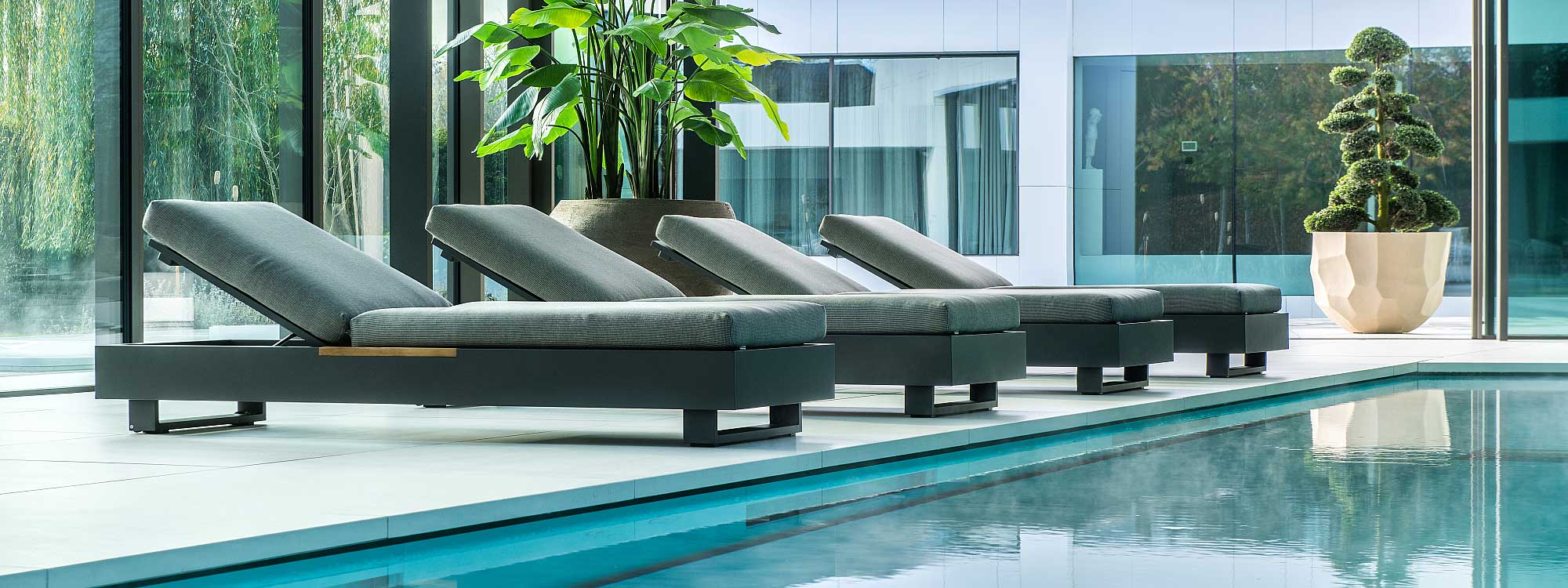 Image of row of 4 Bari luxury sun loungers in charcoal coloured aluminium along interior poolside, with large plant pots and cloud pruned shrub in the background