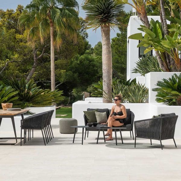 Image of hot white-washed terrace with woman in sunglasses sat on charcoal black-colored Fortuna Rope 2 seat garden sofa