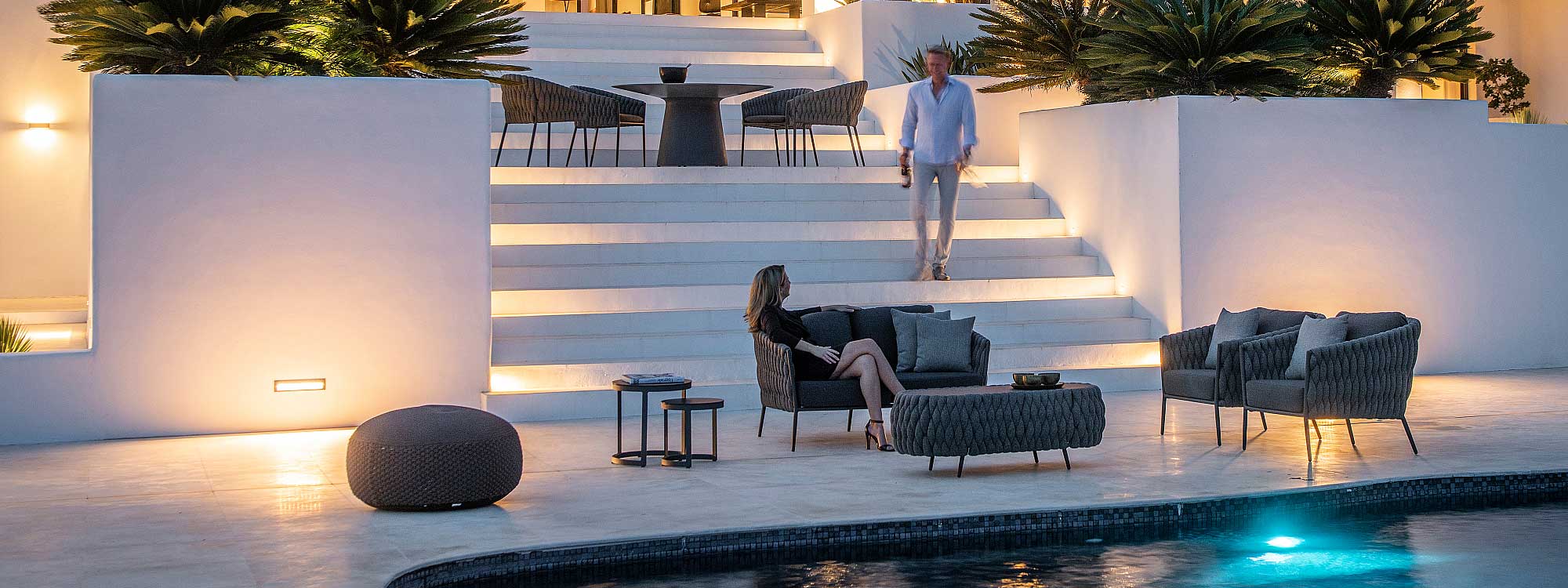 Nighttime image of illuminated white-washed terrace with Fortuna Socks garden sofa and lounge chairs in Charcoal black finish, with Fortuna Socks dining chairs shown at the top of large flight of stairs