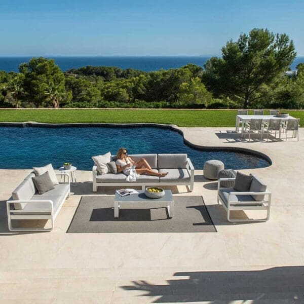 Image of woman lying on Reno modern 3 seat garden sofa by Jati & Kebon, with swimming pool, woodland and sea in the distance beneath blue sky