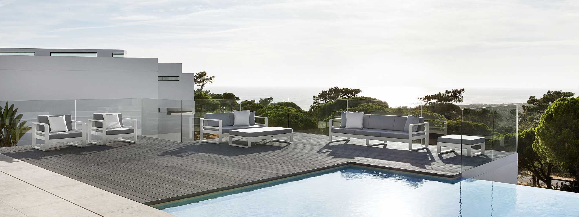 Image of Reno white garden sofas and lounge chairs on a decked poolside, with white-washed building, woodland and sea in the background