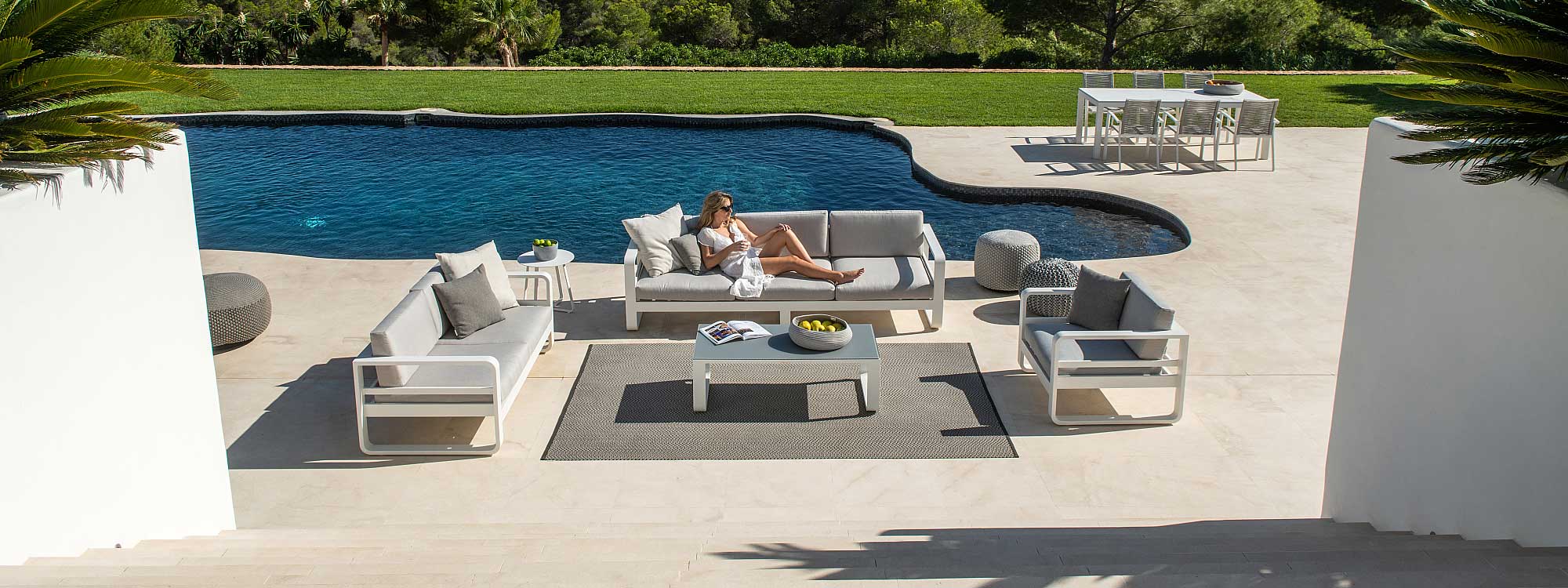 Image of woman lying back in Reno white aluminium garden sofa with grey cushions, shown on poolside surrounded by lawn and white-washed raised flower beds