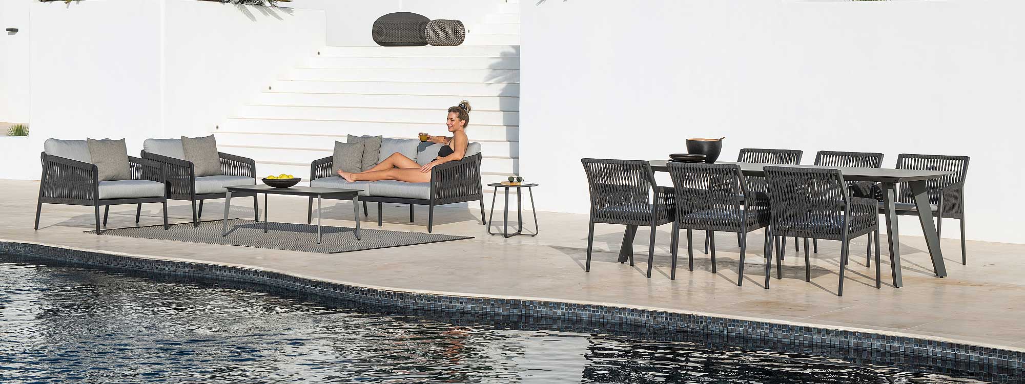 Image of woman lying back on Ritz charcoal coloured garden sofa, next to Ritz outdoor lounge chairs and Ritz modern dining set, shown on sunny poolside against a whitewashed wall.