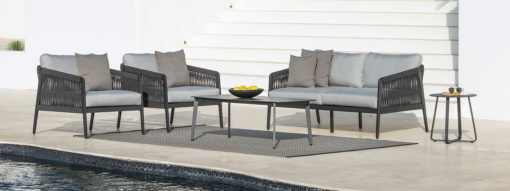 Image of Ritz charcoal coloured garden sofa & lounge chairs with light grey cushions with Ritz coffee table in the centre, shown on sunny poolside with whitewashed flight of stairs and wall in the background