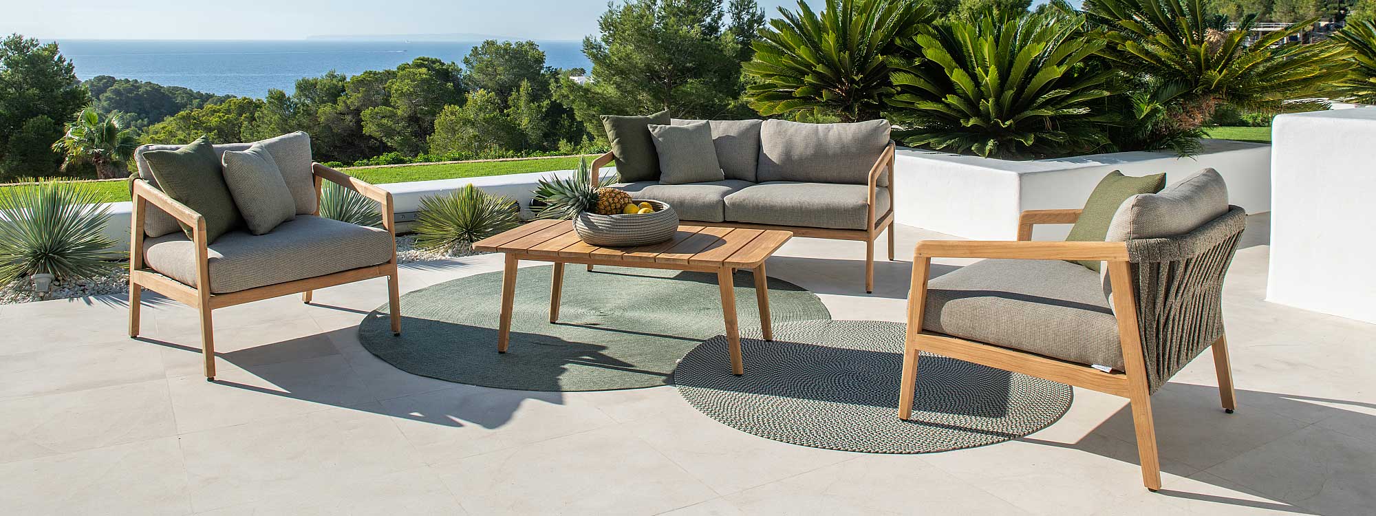 Image of Ritz teak garden sofa and lounge chairs in FSC teak by Jati & Kebon, shown on sunny whitewashed terrace, with lawn, palms, trees and sea in the background