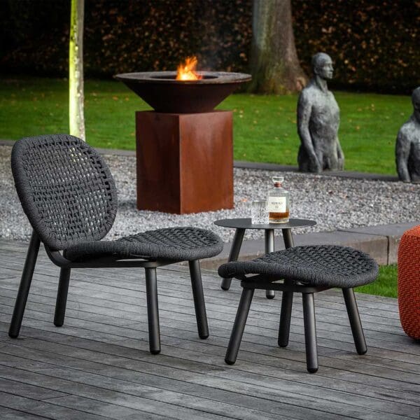 Image of Skate black garden lounge chair and woven foot rest on wooden decking, with sculptures and lawn in the background