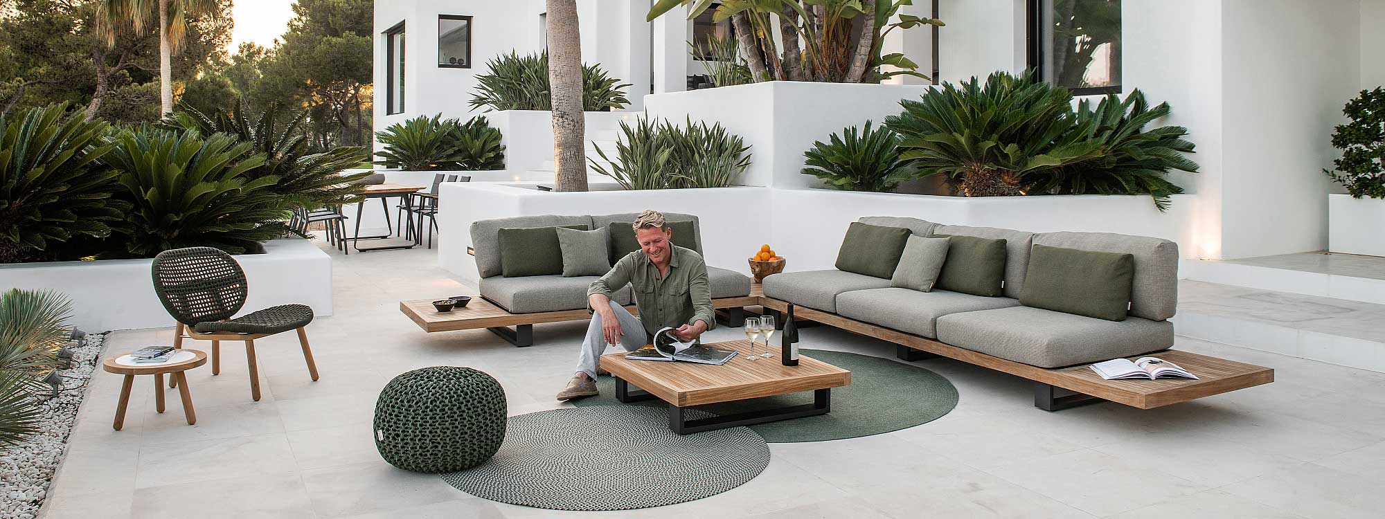 Image of man sat on floor reading a magazine in front of Truro Lounge FSC teak garden sofa, shown on white-washed terrace punctuated by exotic plants