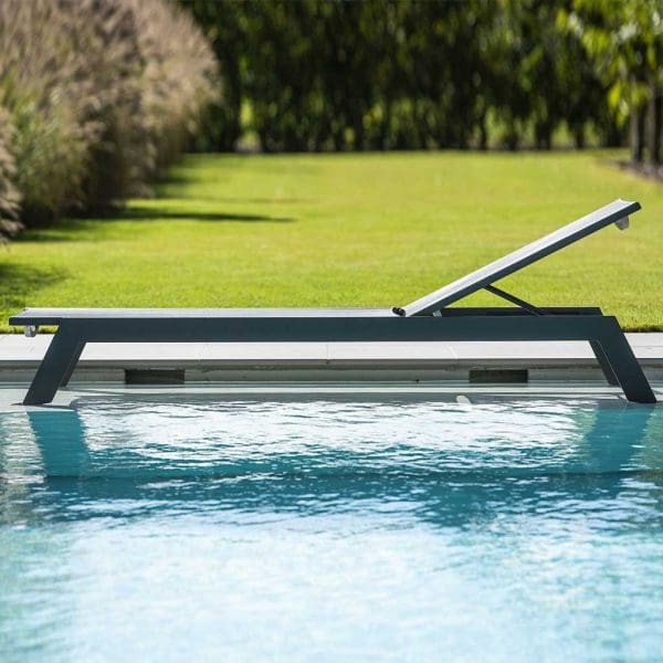 Image of profile of Vigo XL contemporary sun lounger in charcoal colour, shown stood in shallow water in swimming pool with lawn and trees in the background