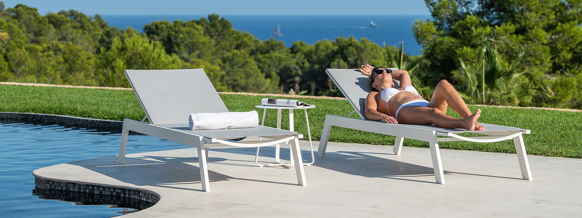 Image of pair of Vigo XL white sun loungers with woman lying in one, with lawns, woodland and blue sea and sky in the background
