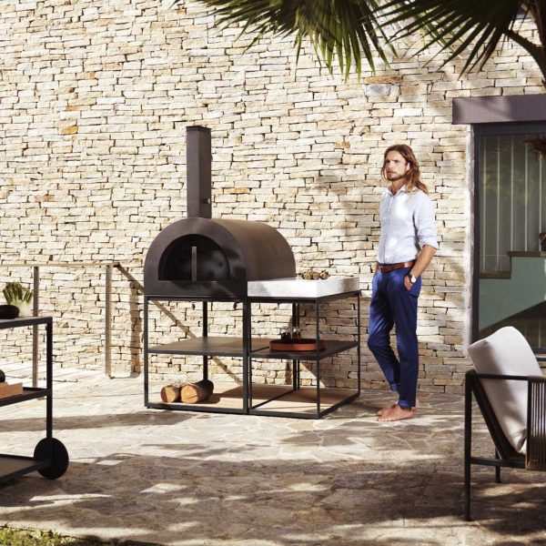 Image of long-haired dude stood next to Roshults minimalist pizza oven and sideboard in anthracite coloured stainless steel, shown in sun and shade of rustic courtyard