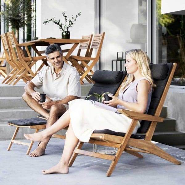 Image of couple sat on Cane-line Flip teak recliner chairs, with Flip folding teak dining furniture in background