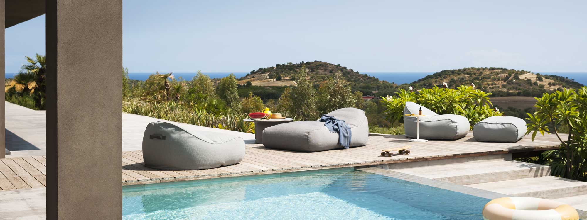 Image of RODA Onda outdoor lounge beanbag and chaise longue bean bag with Apsic low table, shown on sunny poolside decking with undulating Italian countryside in the background