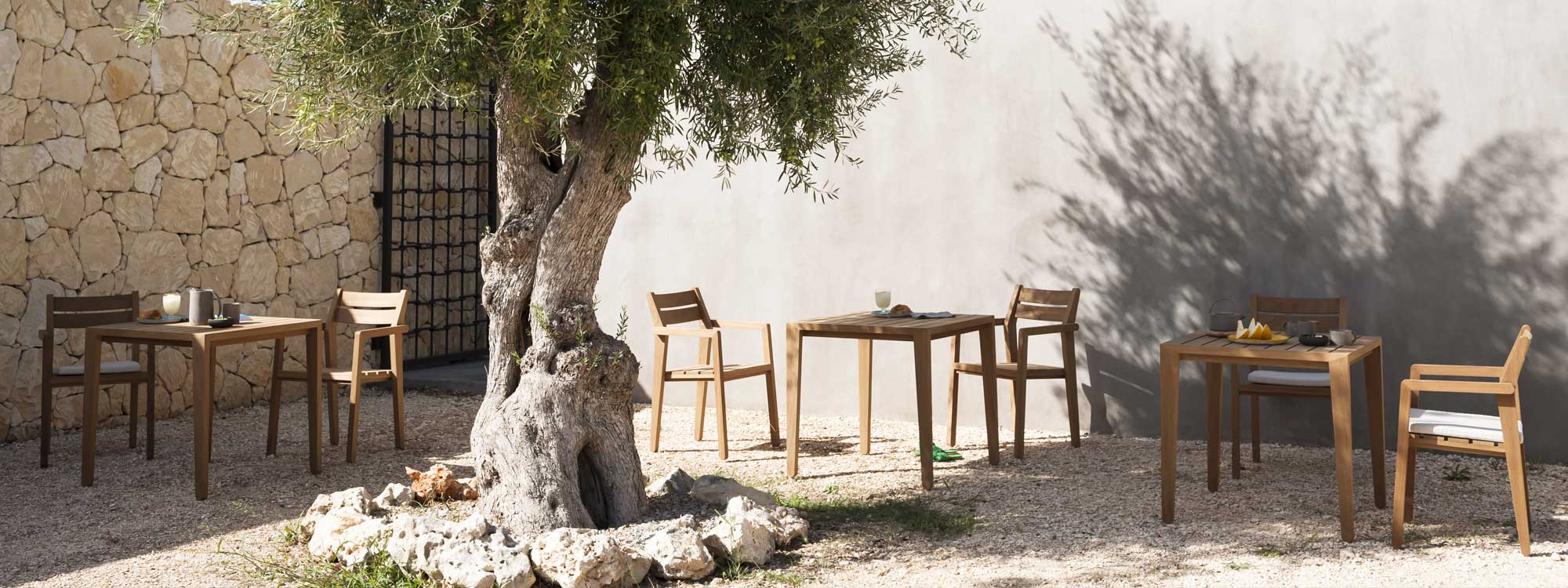 Image of RODA Zania tables and stacking chairs in FSC iroko, shown in sun and shade beneath olive tree in a rural courtyard