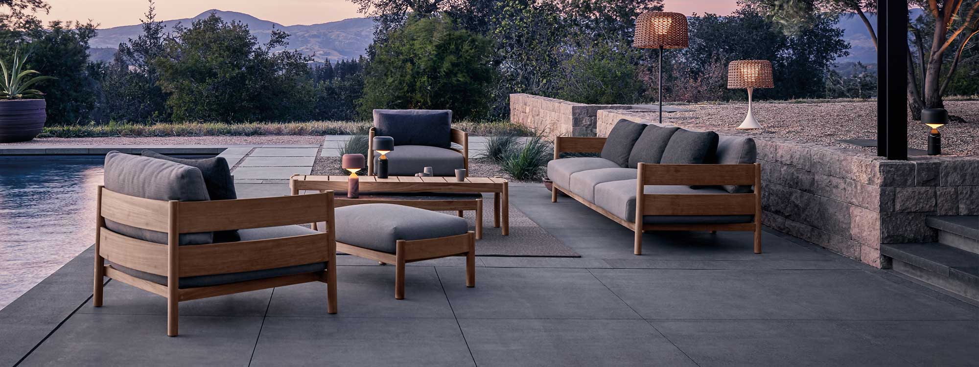 Image at dusk of Haven luxury teak sofa and lounge chairs, together with Mesh outdoor lighting by Gloster, on poolside terrace with trees and hills in the background