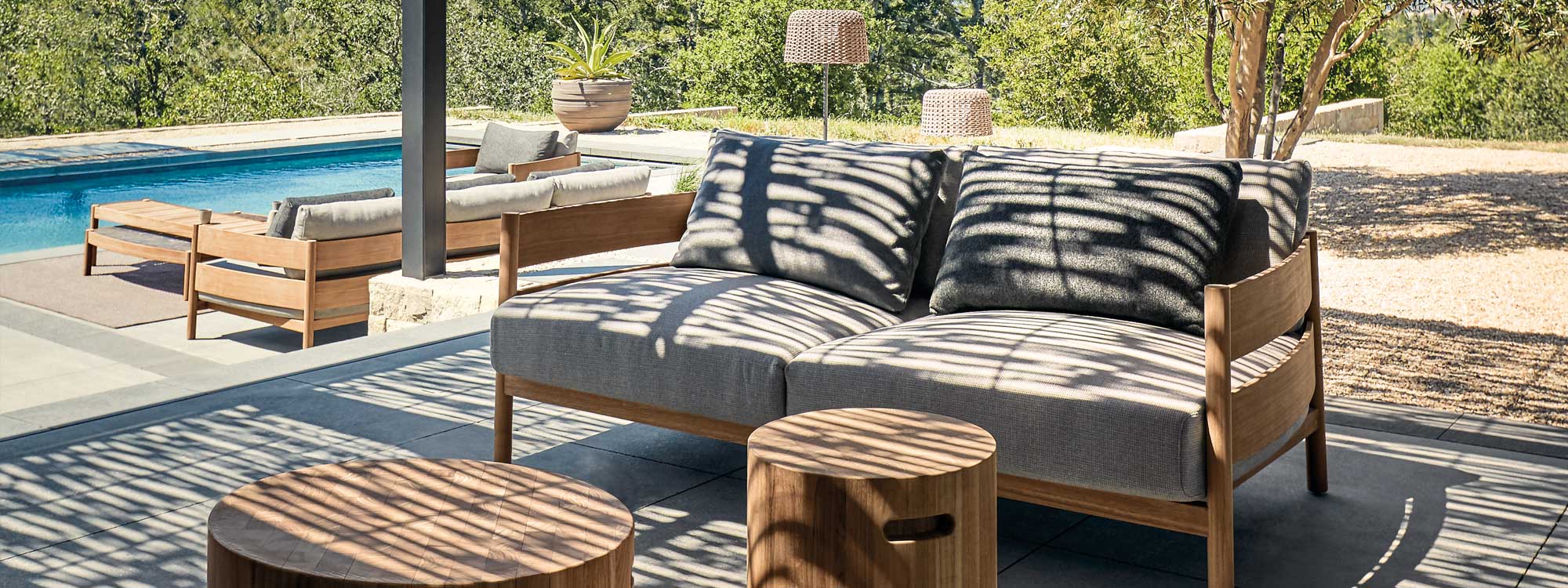 Image of Haven contemporary teak sofas in sun and shade beneath pergola, with more Haven lounge furniture by Gloster in the background next to swimming pool