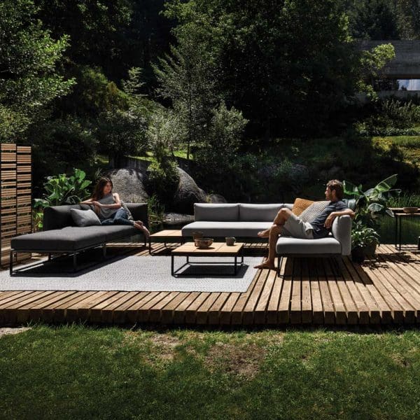 Image of couple relaxing on Maya minimalist garden sofa and daybed by Gloster, shown on wooden decking surrounded by planting