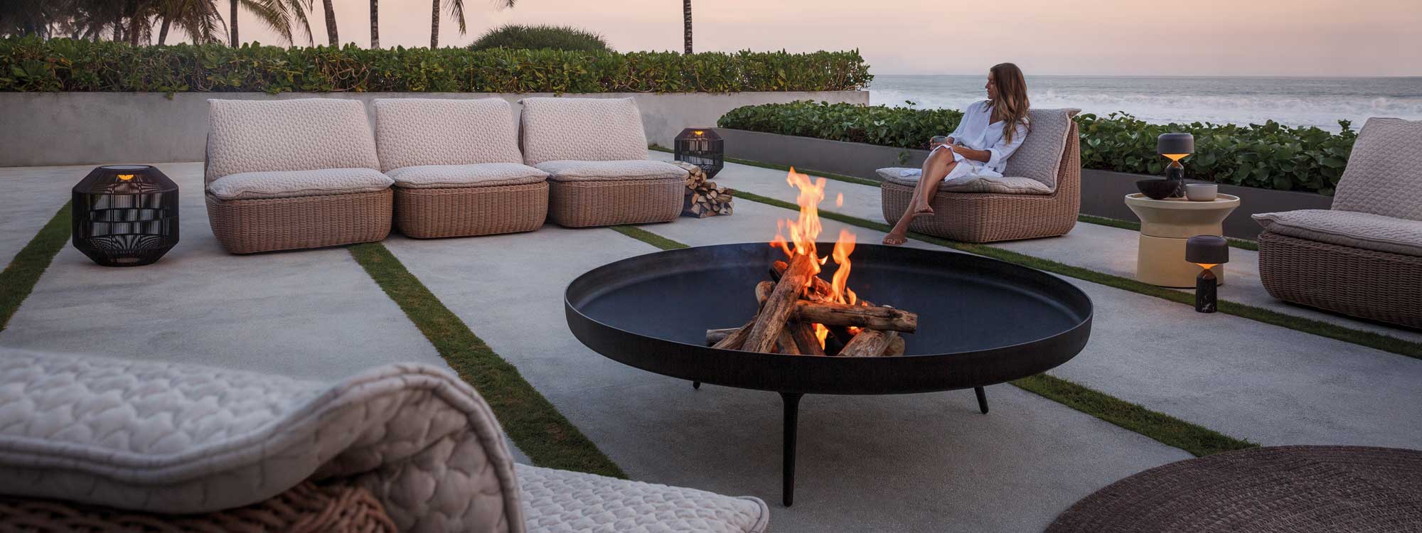 Image at dusk of sleek seaside terrace with woman relaxing on Omada contemporary wicker lounge furniture by Gloster, with roaring fire pit in the centre