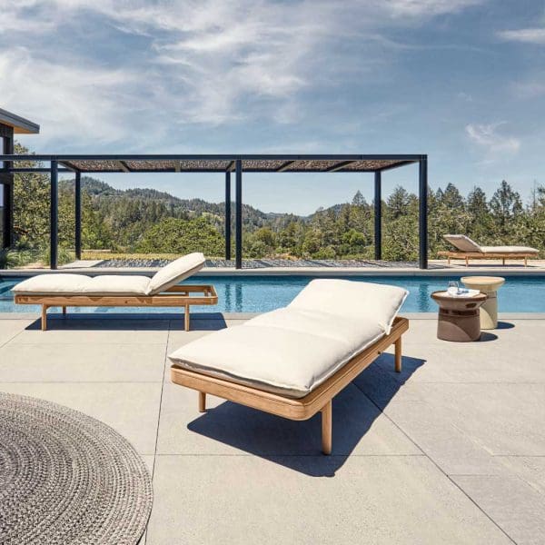 Image of pair of Saranac luxury teak sun loungers by Gloster, shown on sunny terrace with pergola and blue sky and green hills in the background