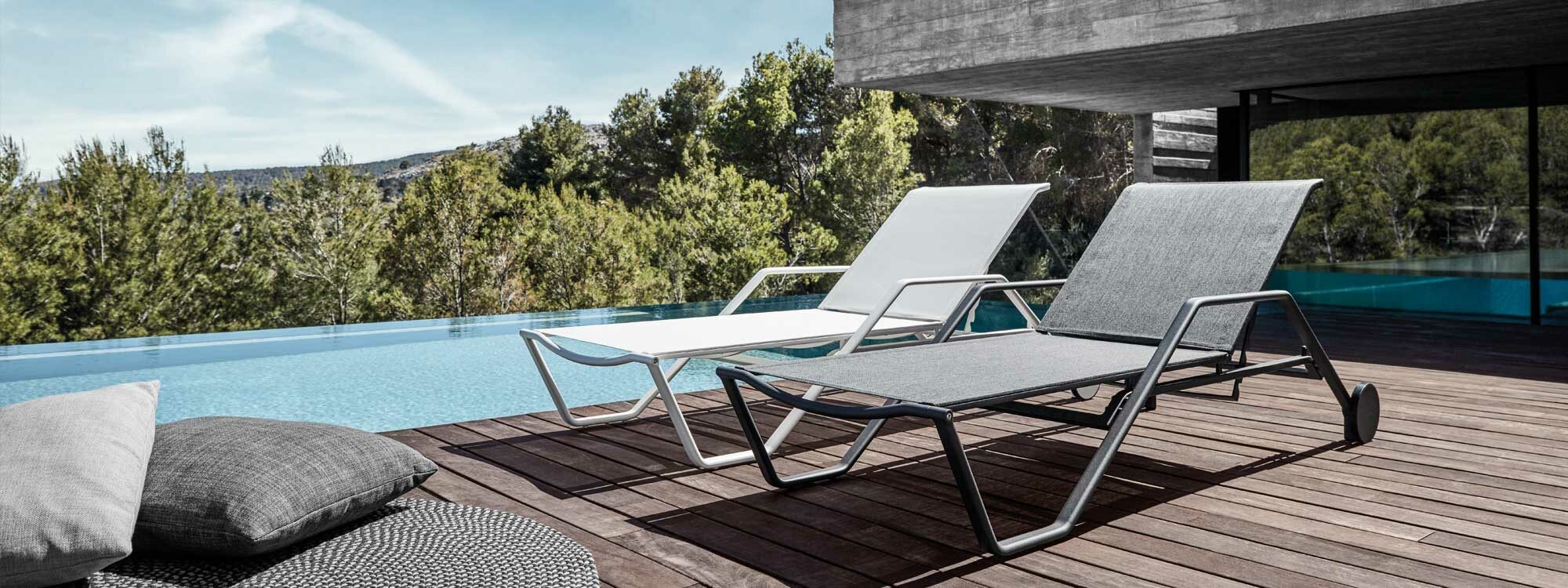 Image of pair of Gloster 180 contemporary sun loungers on decking beside horizon pool with woodland and blue sky in the background