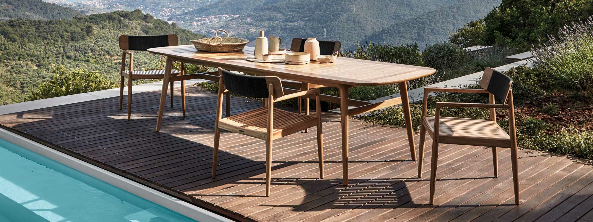 Image of Archi modern teak garden chairs and Gloster teak dining table on sunny poolside, with undulating hills and countryside in the background