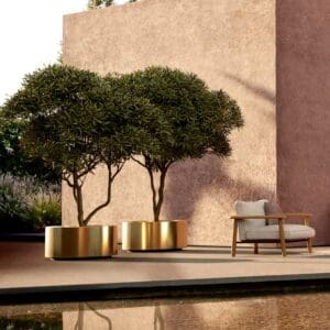 Image of late afternoon sun glinting on Cuprum large brass planters, shown next to Mambo teak lounge chair and tranquil water feature in the foreground