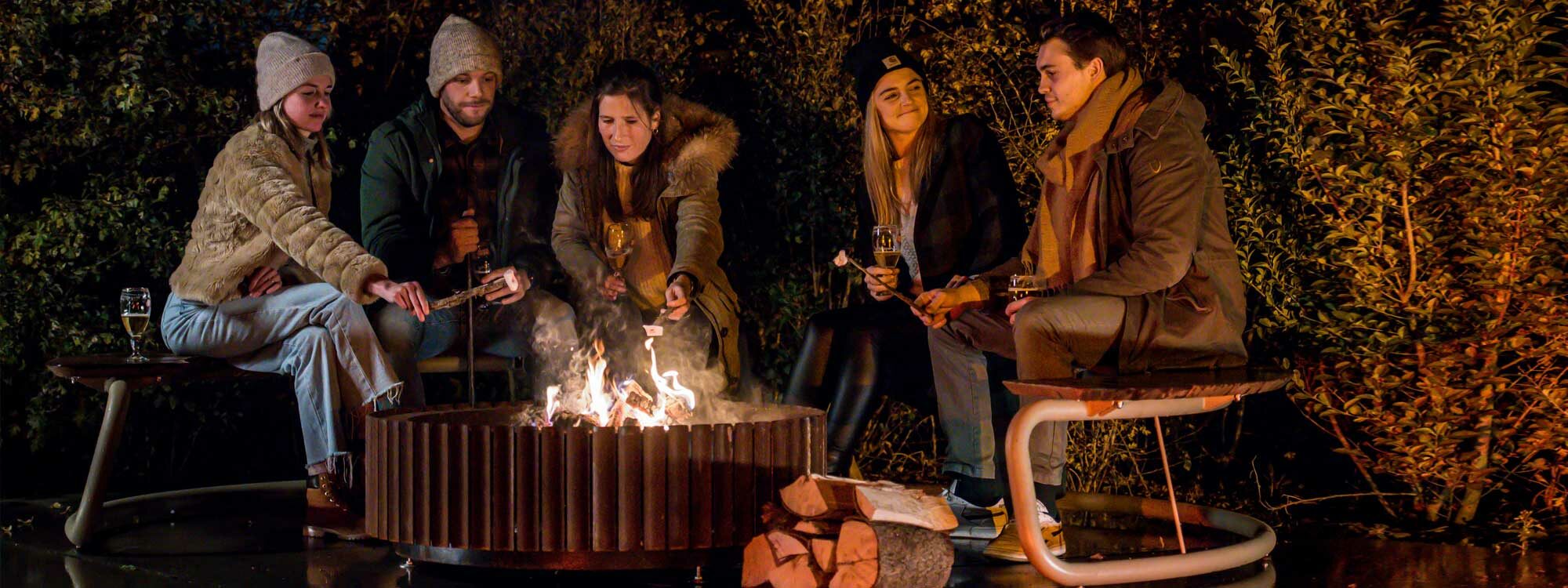 Nighttime shot of friends sat on The Circle round garden bench whilst toasting marshmallows over the Ring corten fire pit by Wünder outdoor furniture