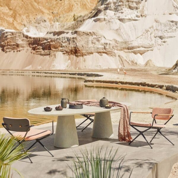 Image of Royal Botania Exes low armchairs and Conix elliptical low dining table on sunny terrace with water and rockface in the background