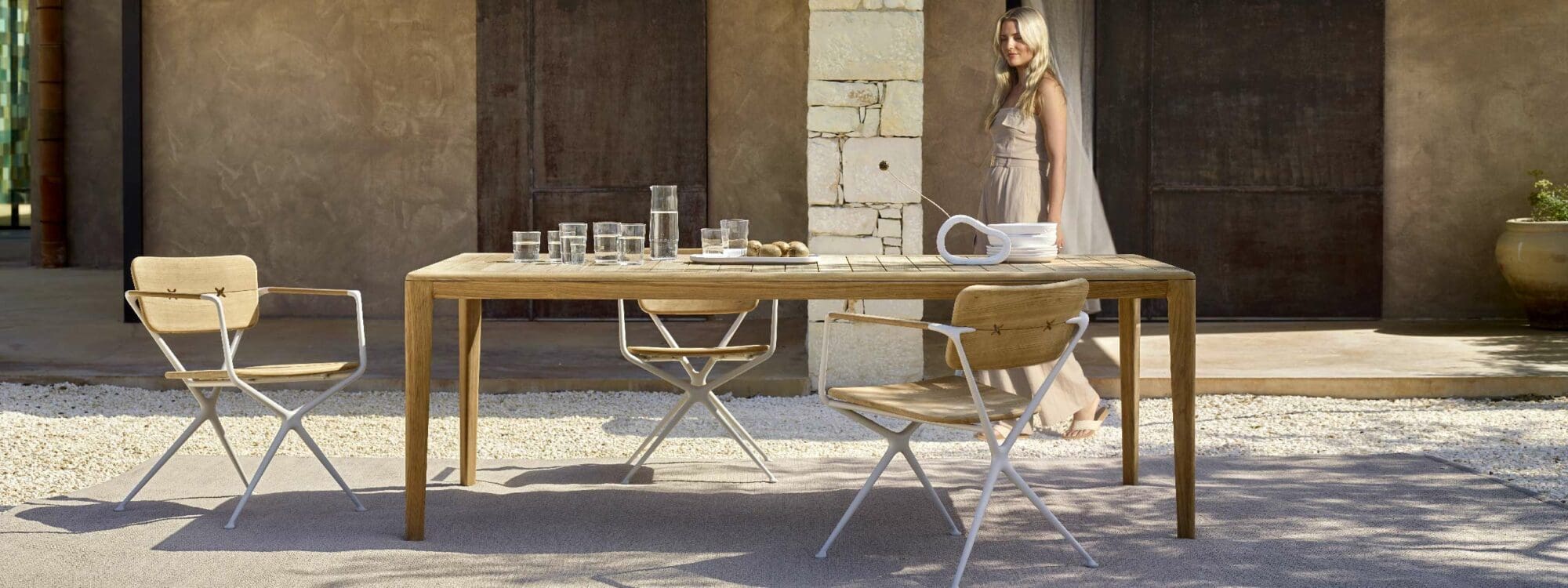 Image of smiling woman walking past Exes garden armchairs and U-NITE teak dining table in a sunny courtyard