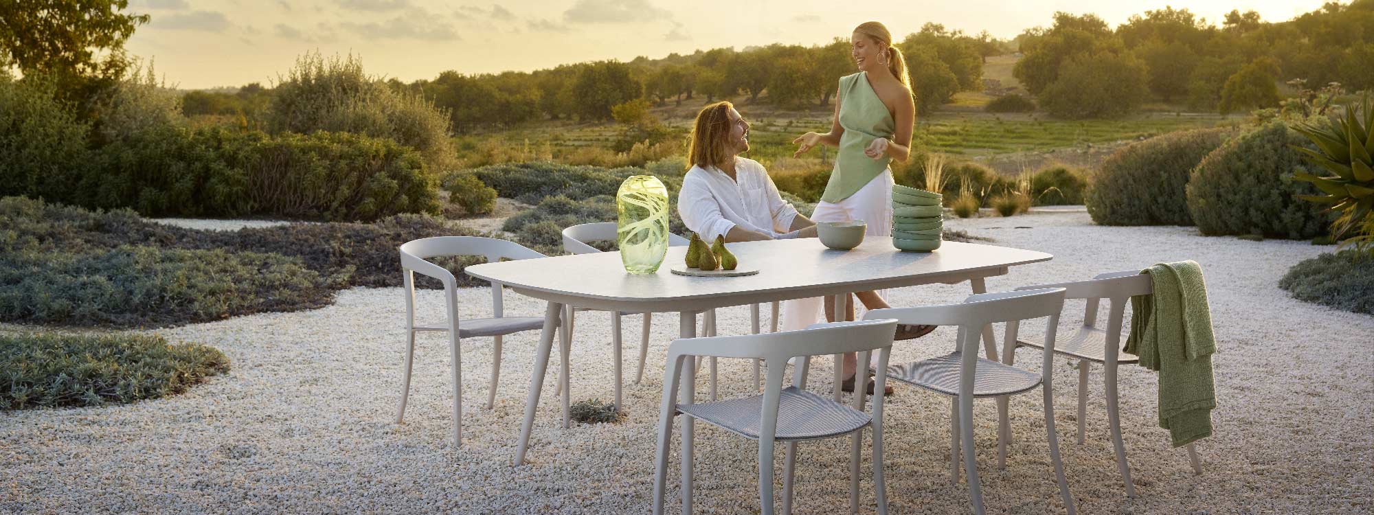 Image of white Styletto garden table and Jive white garden chairs on shingle ground, surrounded by grasses and coastal shrubs