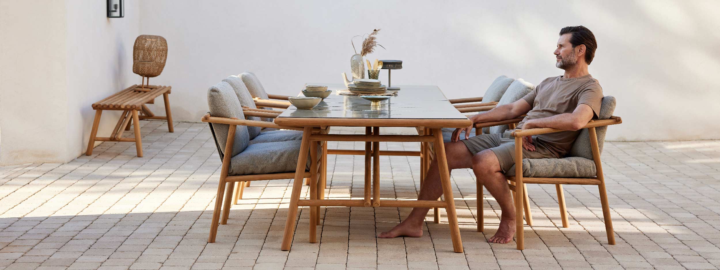 Image of man sat contemplating in Sticks teak garden chair next to Sticks dining table, with Sticks teak bench seat against a wall in the background