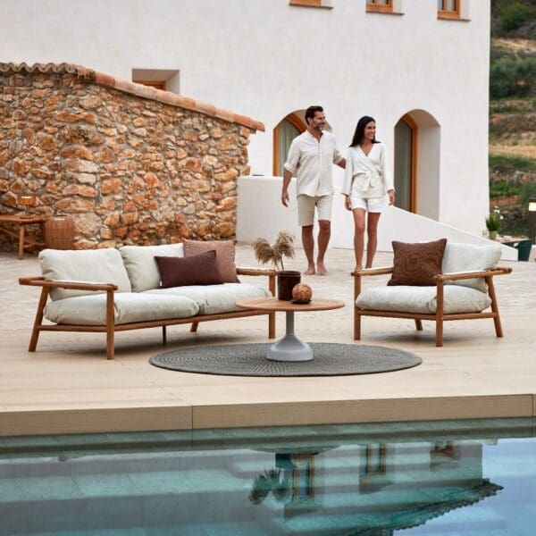 Image of Stick teak sofa and lounge chair on terrace, with still waters of swimming pool in the foreground and smiling couple in the background