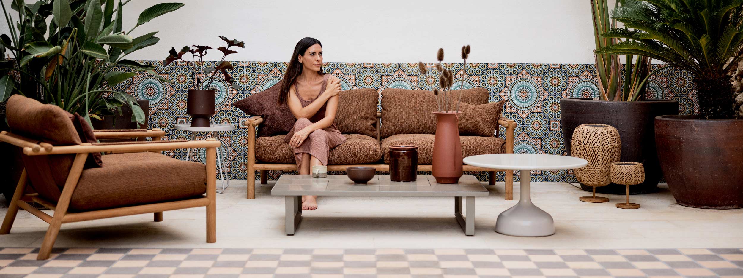 Image of woman sat in Sticks teak garden sofa with Umber Brown cushions, with colourful Moroccan tiles in the background