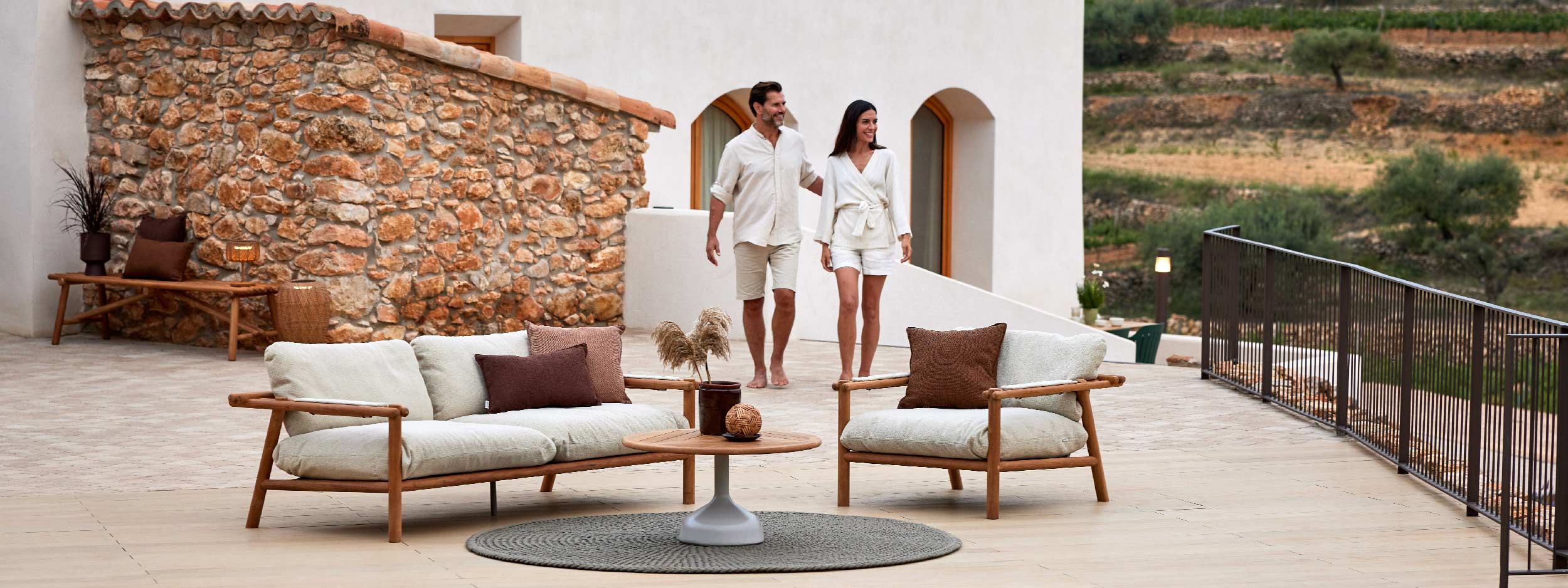 Image of smiling couple walking along terrace next to Cane-line Sticks teak garden sofa and lounge chair