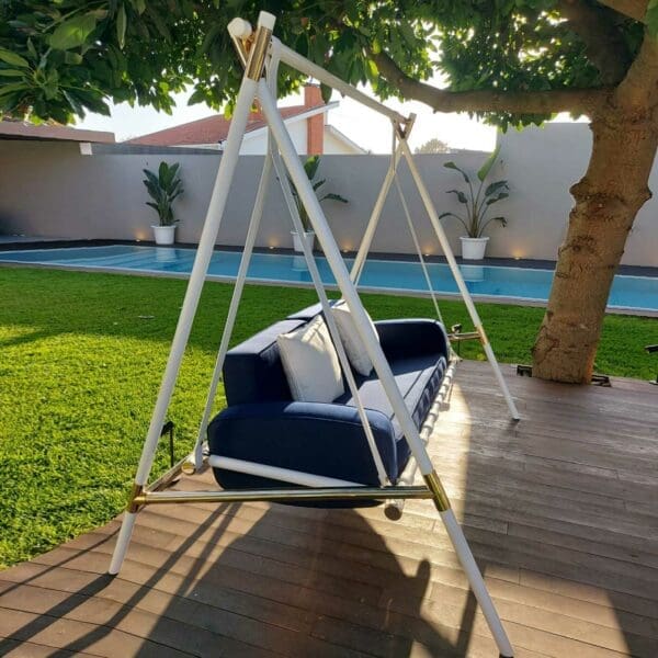 Image of side view of Fable 2 seat sofa swing with white frame and navy blue cushions, on decked platform with tree and swimming pool in the background
